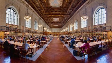 Readers study quietly at long wooden tables inside a bright public library reading room.