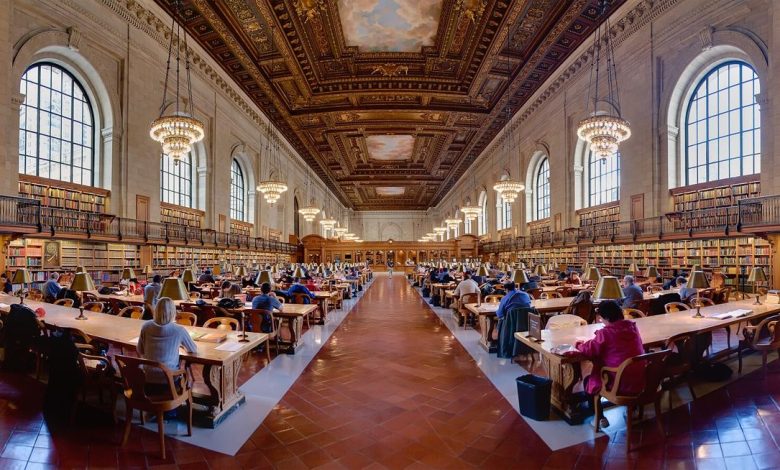 Readers study quietly at long wooden tables inside a bright public library reading room.
